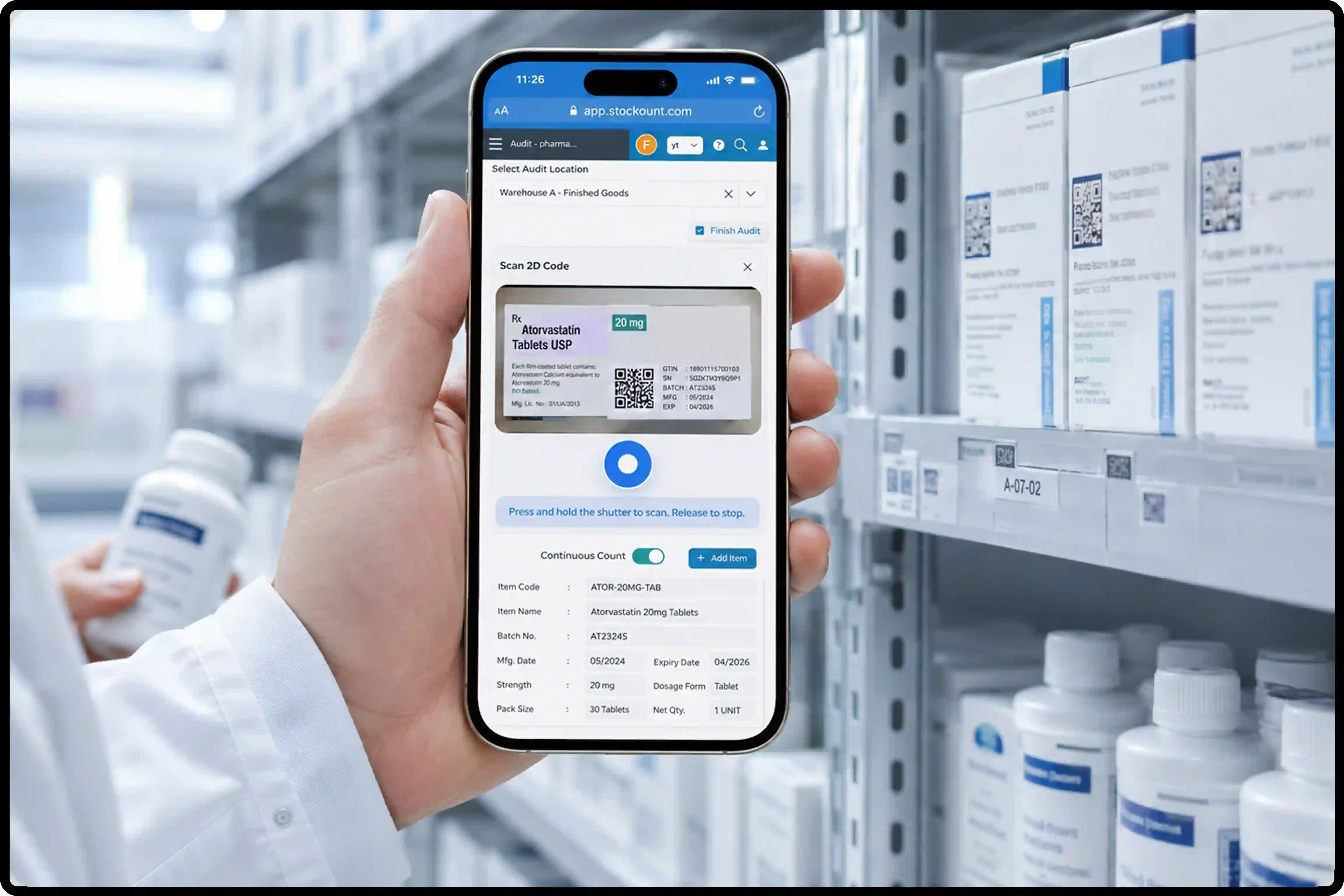 Hand holding a smartphone scanning a QR code on a pharmaceutical product label in a warehouse inventory management system, with medicine boxes and bottles on storage shelves in the background.